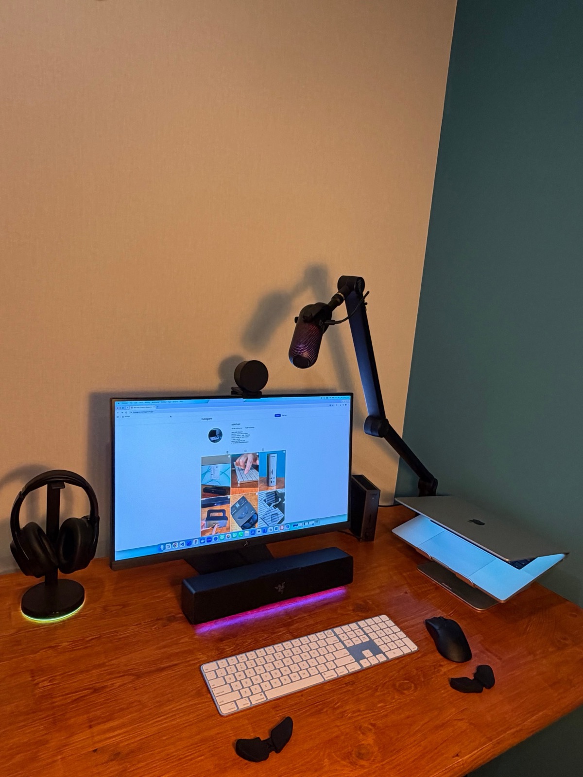 My desk from an angle: a monitor showing my Instagram on a honey-wood desk against a teal wall, a Razer soundbar glowing pink underneath, a white Magic Keyboard, a MacBook Pro on a stand and a boom mic.